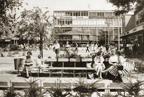 Queens Square. People sitting on benches. Bandstand in the background.