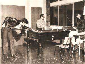 Three young men playing bar billiards. Two are wearing leather jackets.