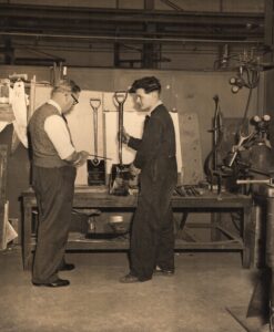 Black and white image of two men with spades standing up on a work bench.