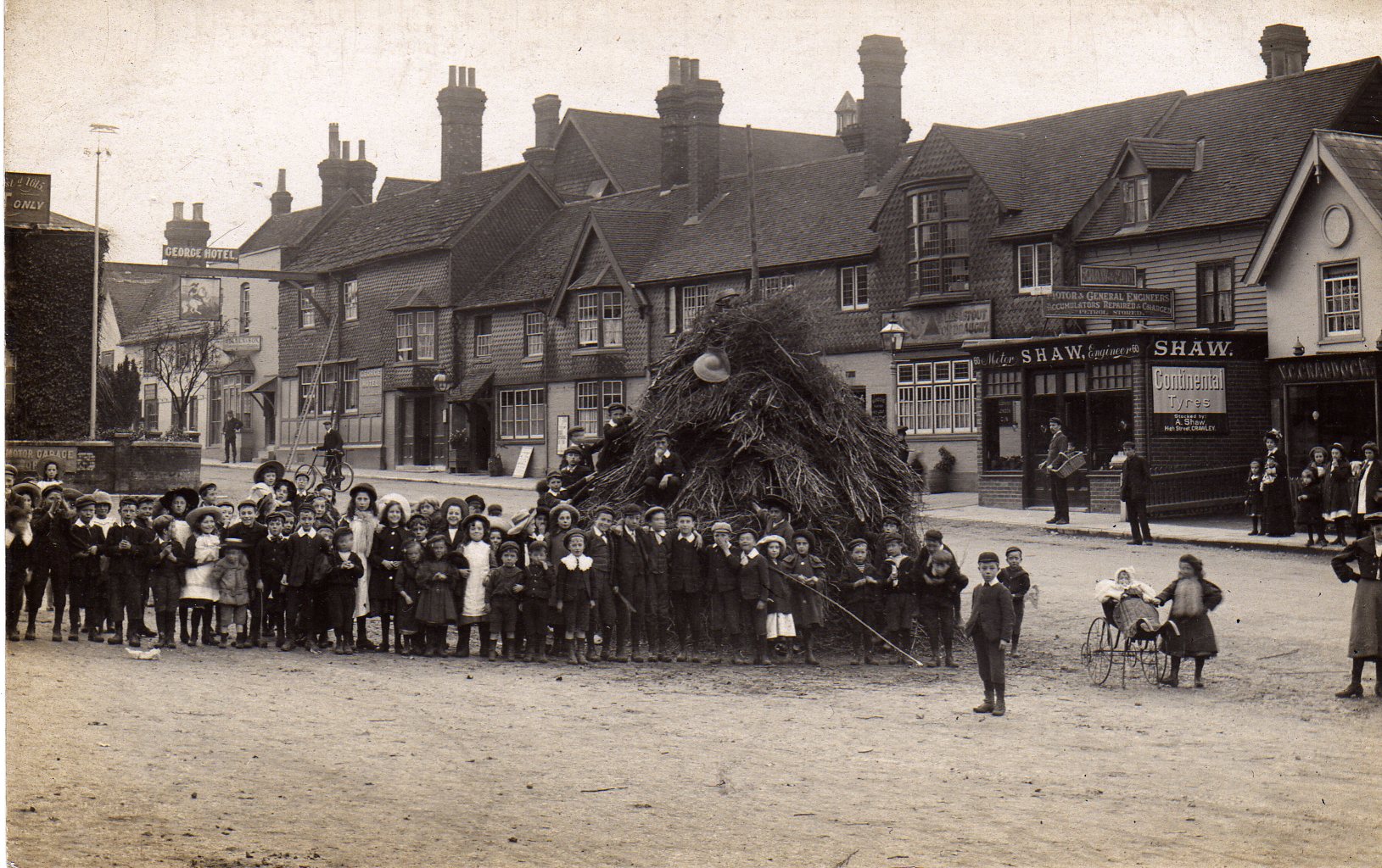 Black and white photograph of a large group of children in front of an unlit bonfire in the street, buildings behind them. 