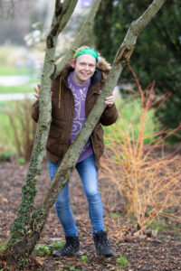 Colour photograph of a man in dark green coat standing behind a tree.