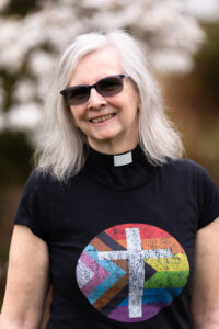 Colour image of a woman with grey hair wearing a clerical collar, dark t-shirt with pride logo on.