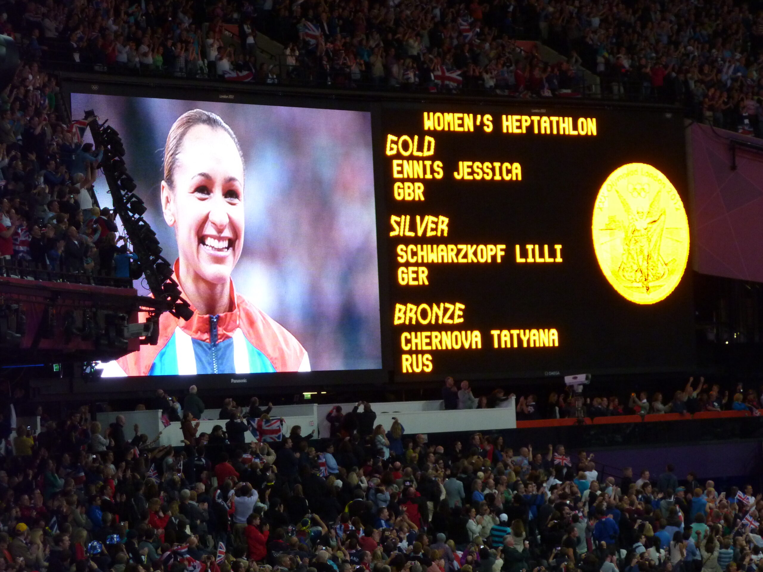 Photograph of a large screen. Left side has the image of a woman and right side has the image of a gold medal with text. 