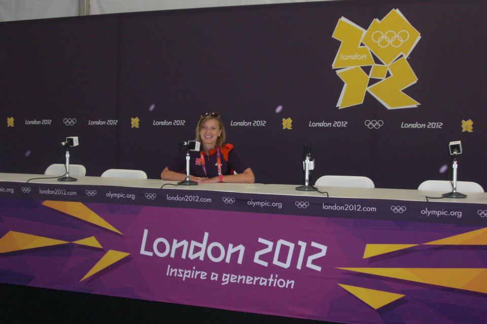 Woman sits at a table, with purple "London 2012" banner on the front. 