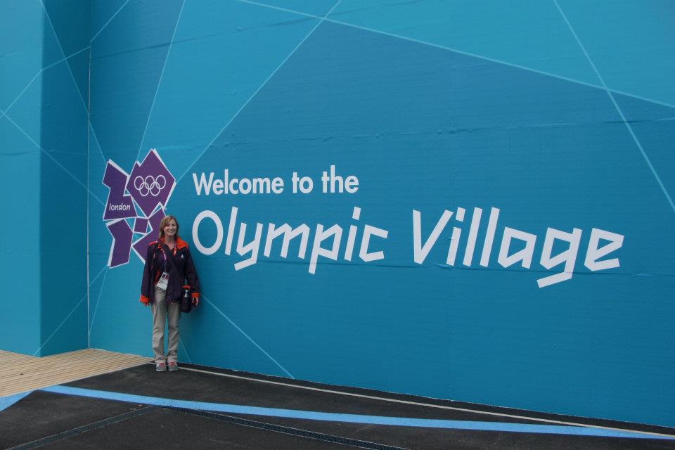 Woman standing in front of blue wall, on the wall in white "Welcome to the Olympic Village" is written