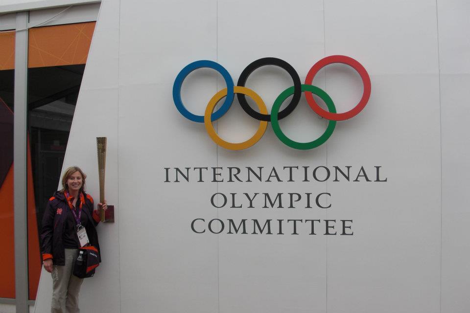 Woman standing holding gold object in front of white wall which has the Olympic rings on the side and "International Olympic Committee" below. 