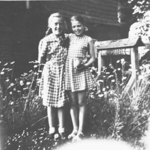 Black and white photograph of two girls in dresses standing next to each other. There is long grass around them and a building behind. 