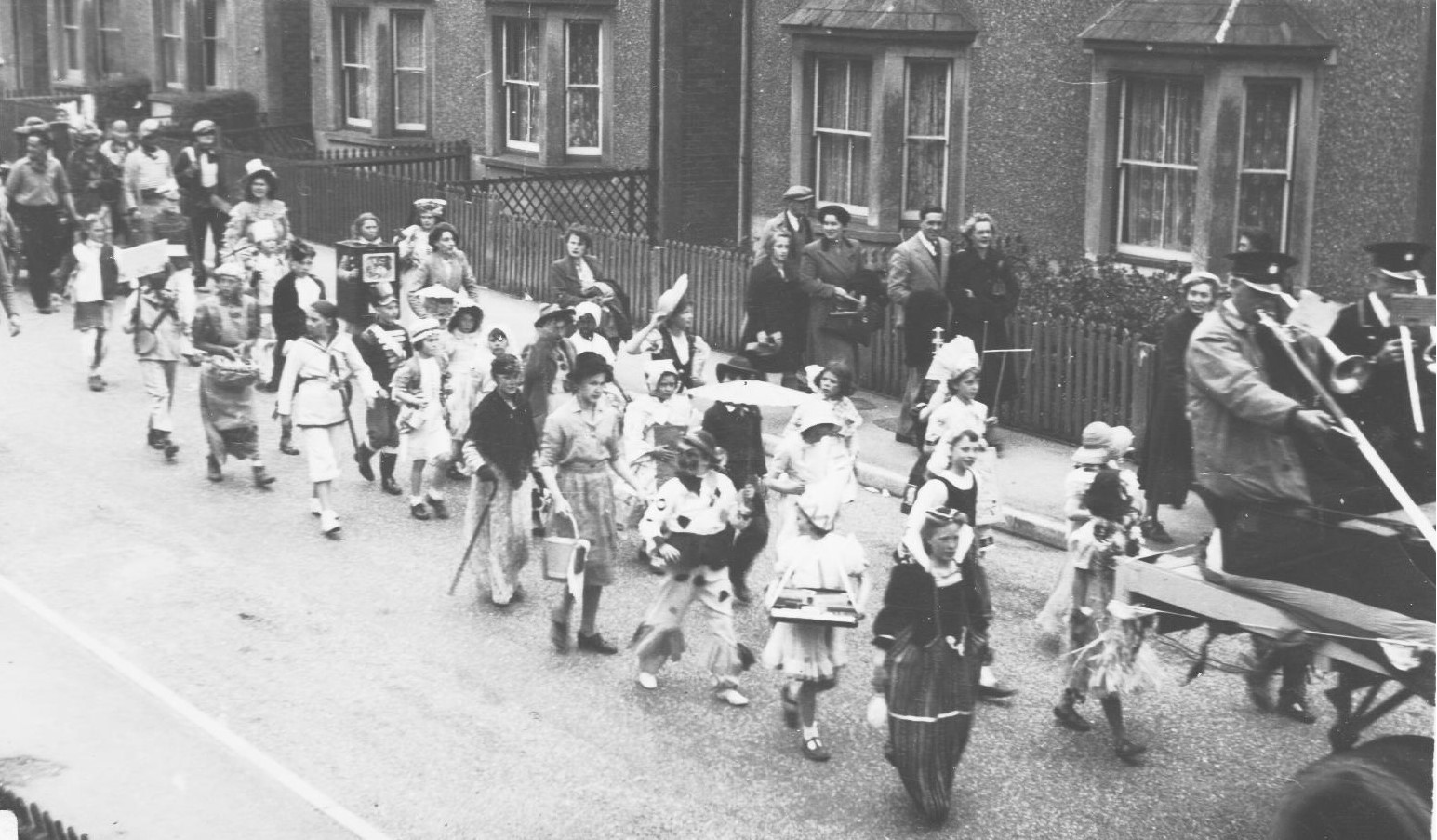 Black and white photograph of a children's parade though a row of houses. The children are dressed up. 