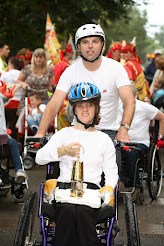 Woman in a wheel chair with someone pushing behind, both are wearing white. Woman in the wheel chair is holding a gold lantern. 