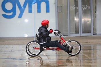 Person in a hand powered three wheel bike passes a building behind them. 