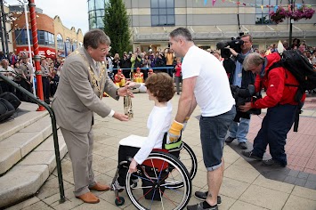 Woman in a wheel chair wearing white hands a gold lantern to a man standing in a suit.