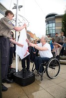 Person in a wheel chair is raising a while flag up a flag pole. Aided by a person in brown shirt and blue trousers. 