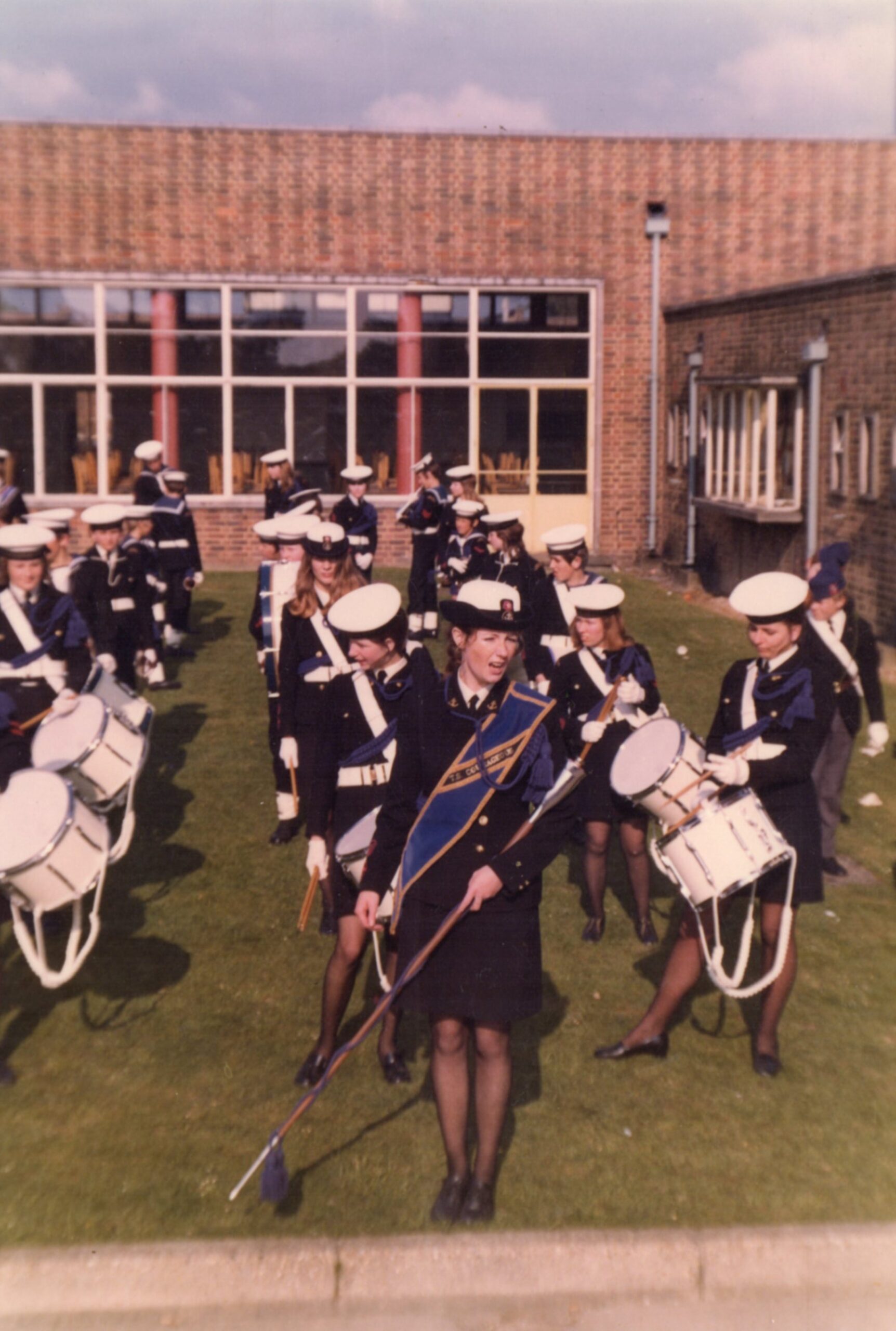 Colour photograph of a marching band standing on a field with a building behind them. 