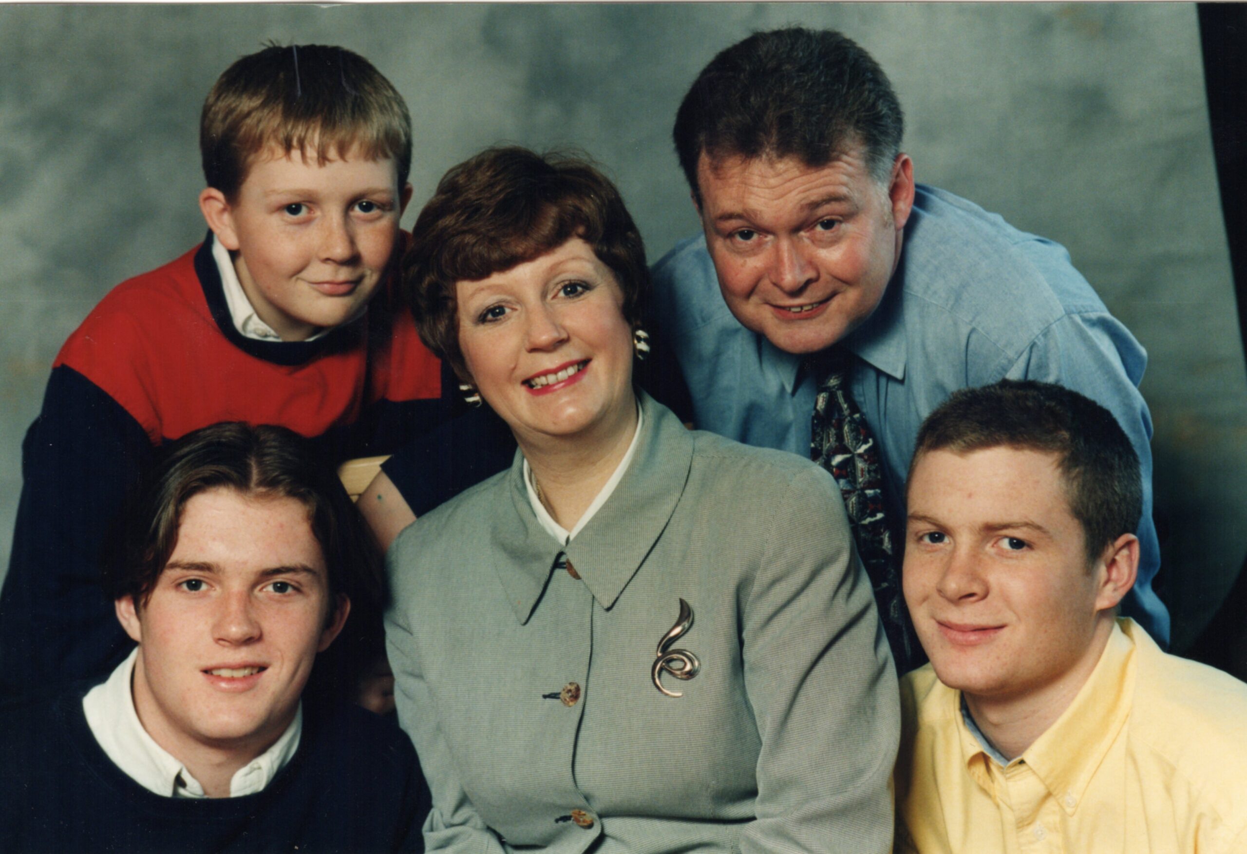 family photo, the only woman is sitting in the middle with a son sitting each side of her, another son and husband behind her. 